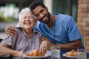 A male caregiver is helping an elderly woman, and they are enjoying some time together by eating